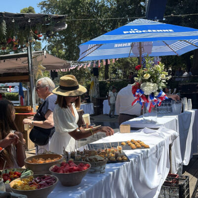 Woman in a white dress and straw hat serves bite-sized snacks at an outdoor market, with bowls of fruit on a long, white-draped table under a blue umbrella.