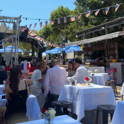 Outdoor social event with guests seated at white-clothed tables, small floral centerpieces, and string lights under patriotic bunting and flags, sunny day.