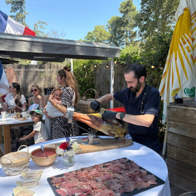 Outdoor backyard party: a chef carves a ham leg on a stand while guests enjoy snacks at a long table under blue skies.